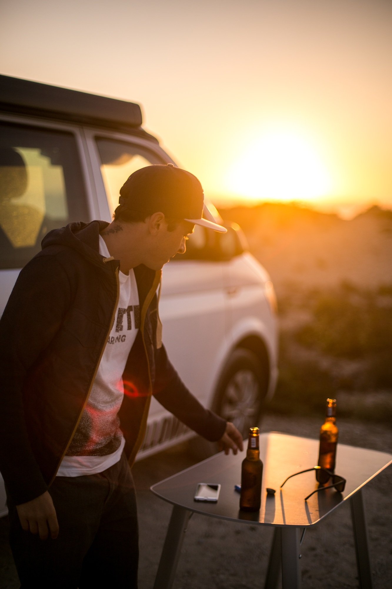 A man grabs a beer in front of the sun setting over the ocean during a road trip in a campervan
